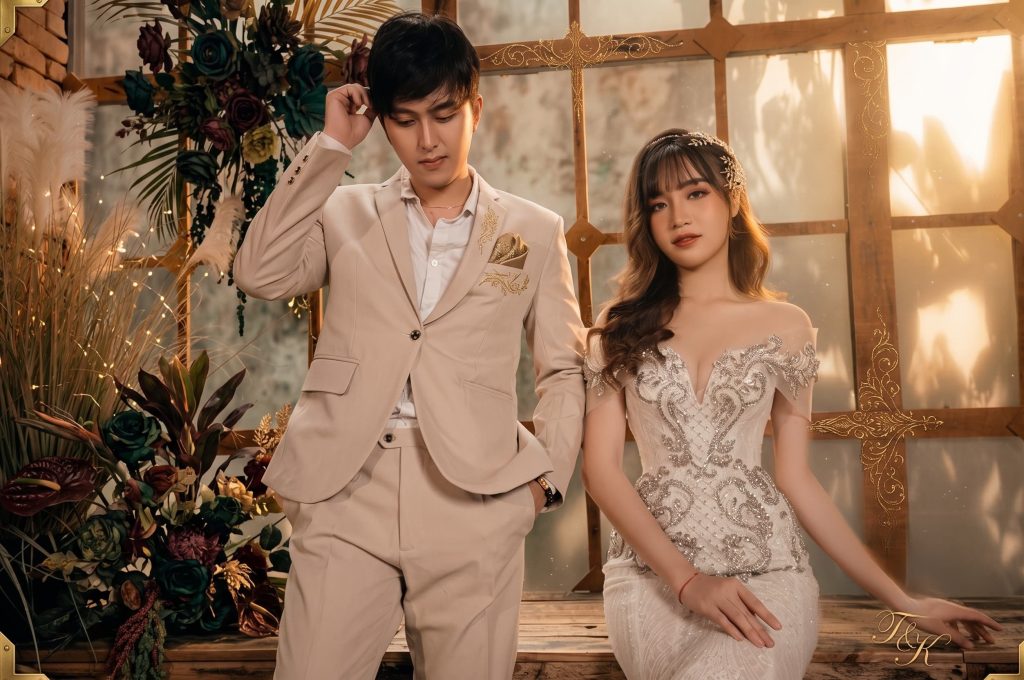 Asian bride in a beaded off-the-shoulder gown and groom in a beige suit posing against a warm floral backdrop.