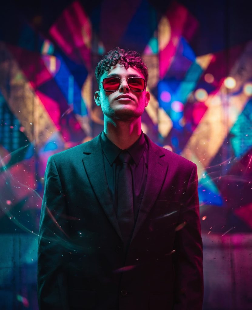 Young man in a black suit, black tie, and red-tinted sunglasses posing in front of a colorful neon background.