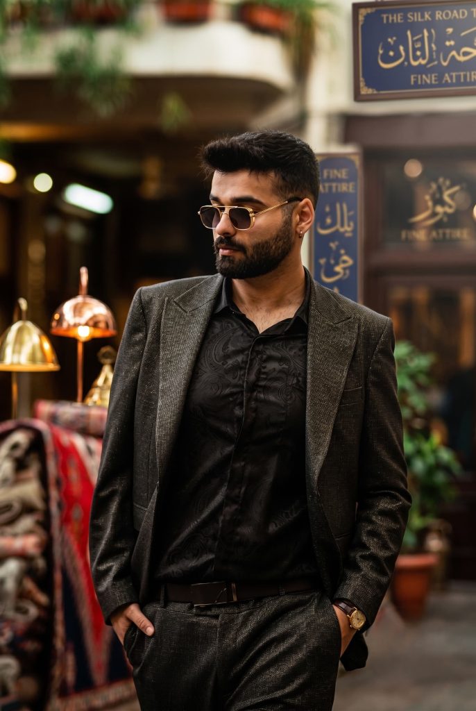 Bearded man in a textured dark suit, patterned shirt, and sunglasses walking through a traditional market square.