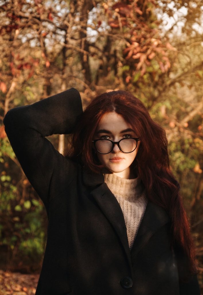 Woman with long auburn hair and glasses wearing a black coat and beige turtleneck sweater in an autumn forest.