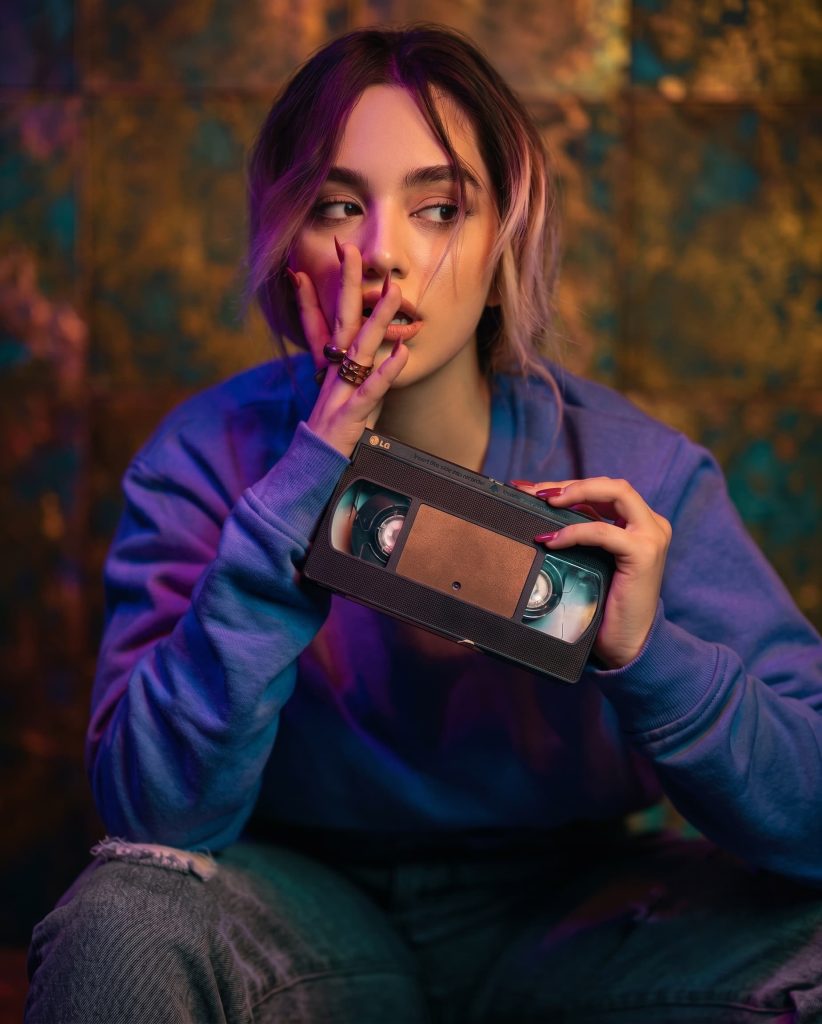Woman holding a vintage VHS tape in a neon-lit room with moody purple lighting.