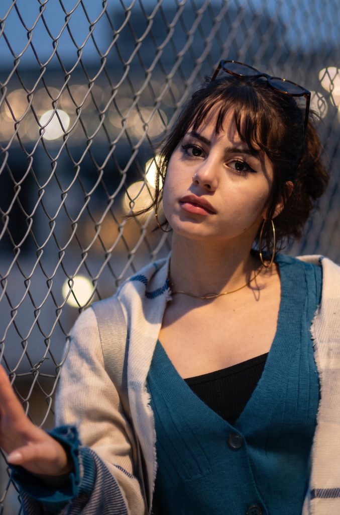 Young woman with dark bangs wearing a teal cardigan and hoop earrings, leaning against a chain-link fence at dusk.