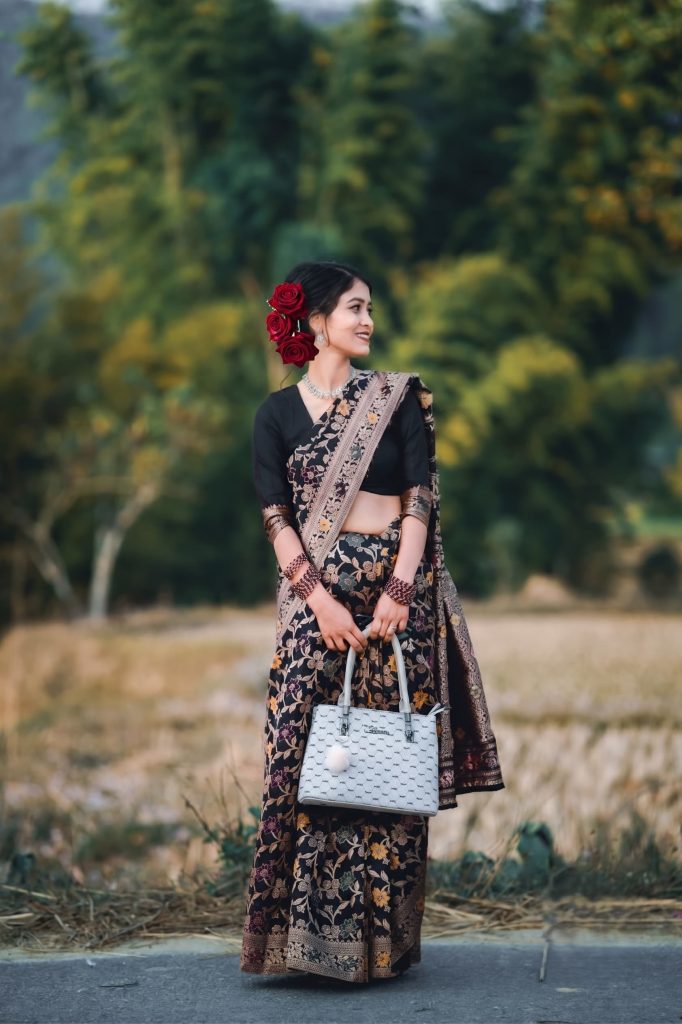 Woman wearing a black floral saree with red roses in her hair, holding a light grey handbag outdoors.
