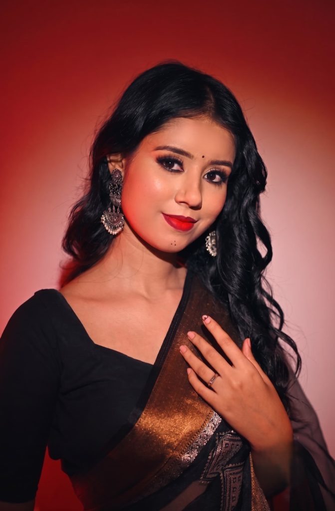 Woman wearing a black saree with a golden border and oxidized jhumka earrings against a red gradient background.