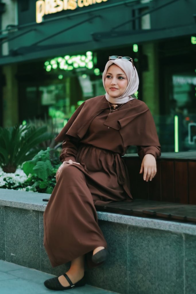 Woman wearing a long brown dress and light-colored hijab sitting outdoors on a ledge near a storefront.