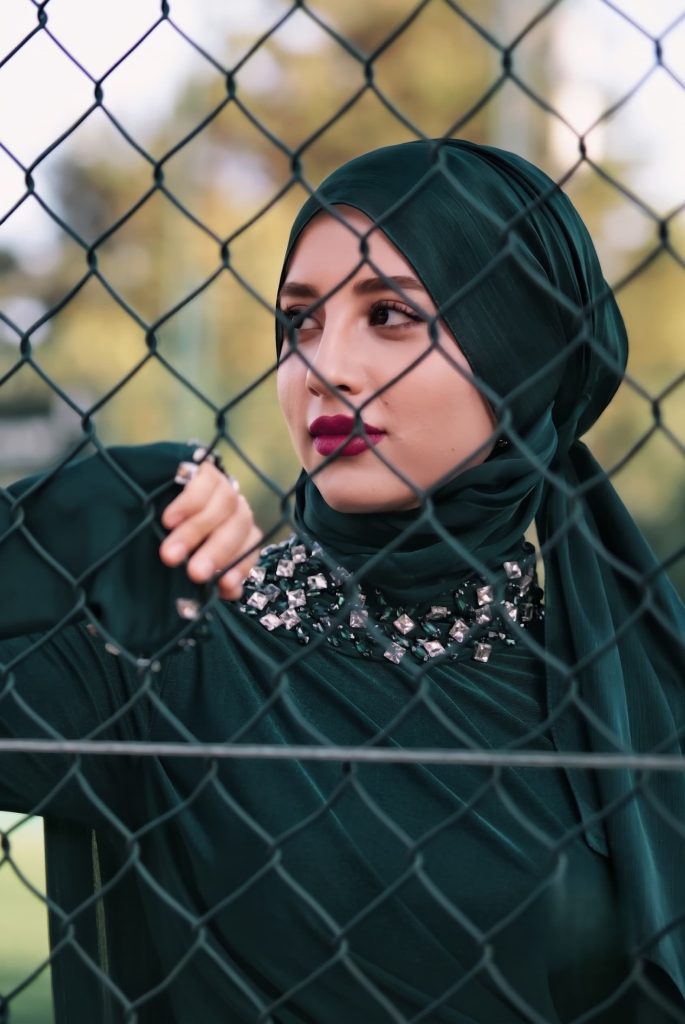 Woman wearing a dark green embellished hijab and dress holding onto a chain-link fence looking away.
