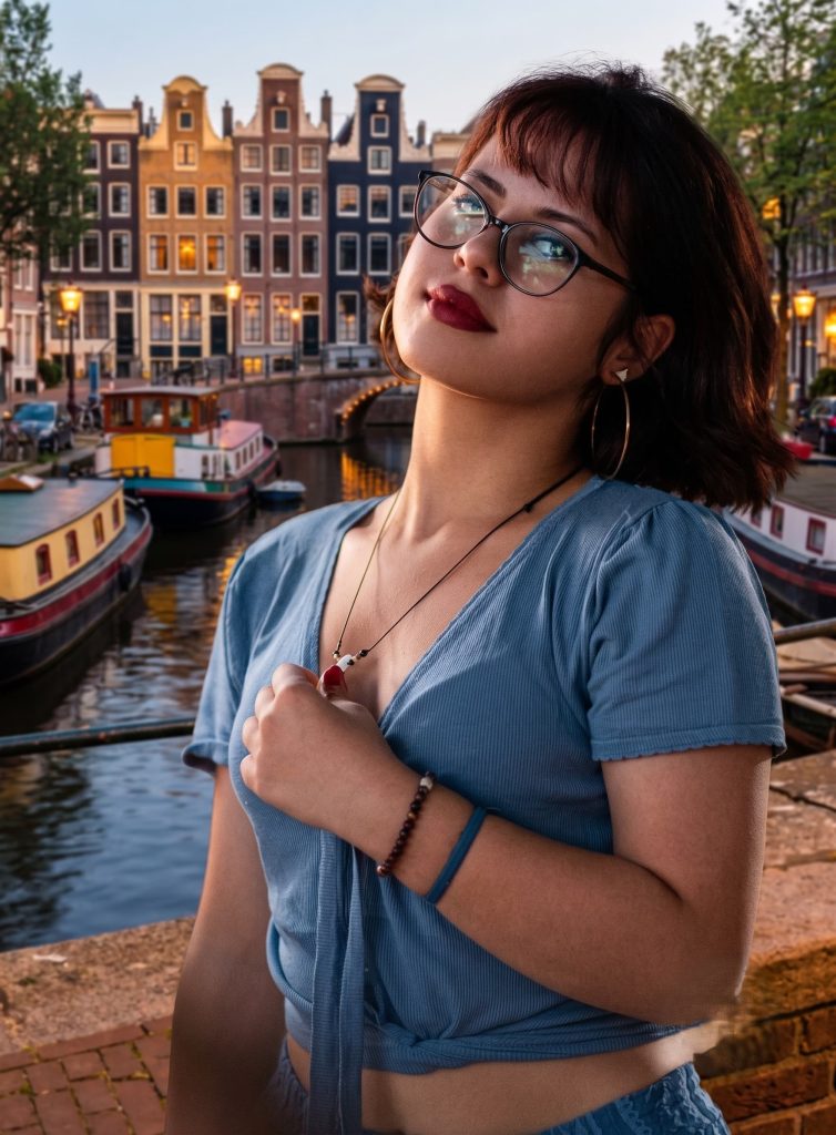 Woman with glasses and red lipstick wearing a blue crop top by an Amsterdam canal with canal houses in the background.