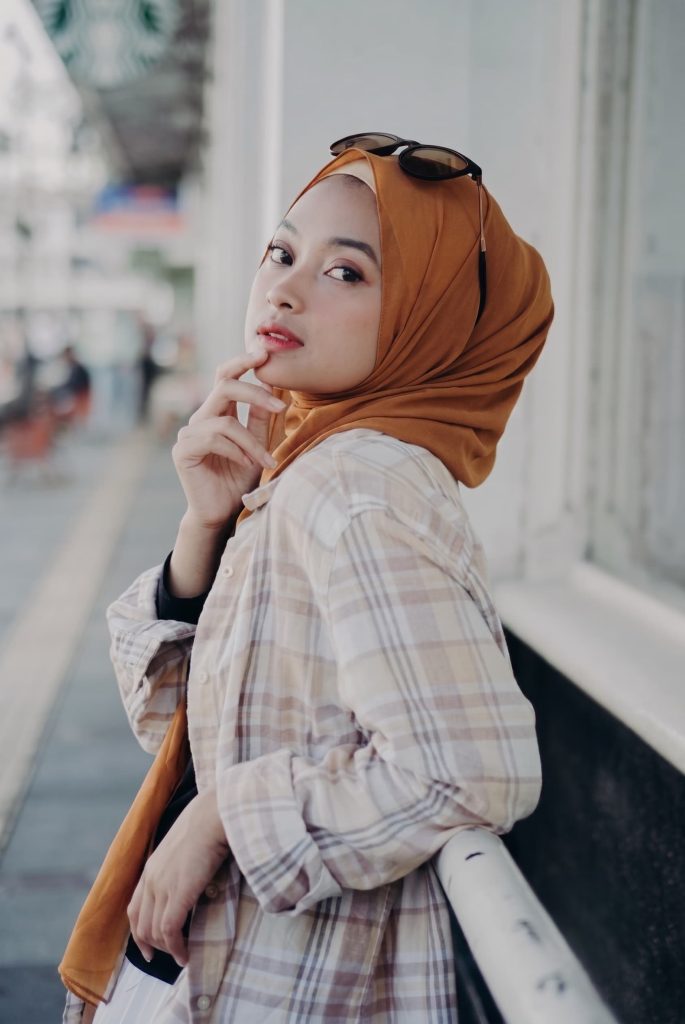 Young woman wearing a mustard yellow hijab, sunglasses on her head, and a plaid button-up shirt outdoors.