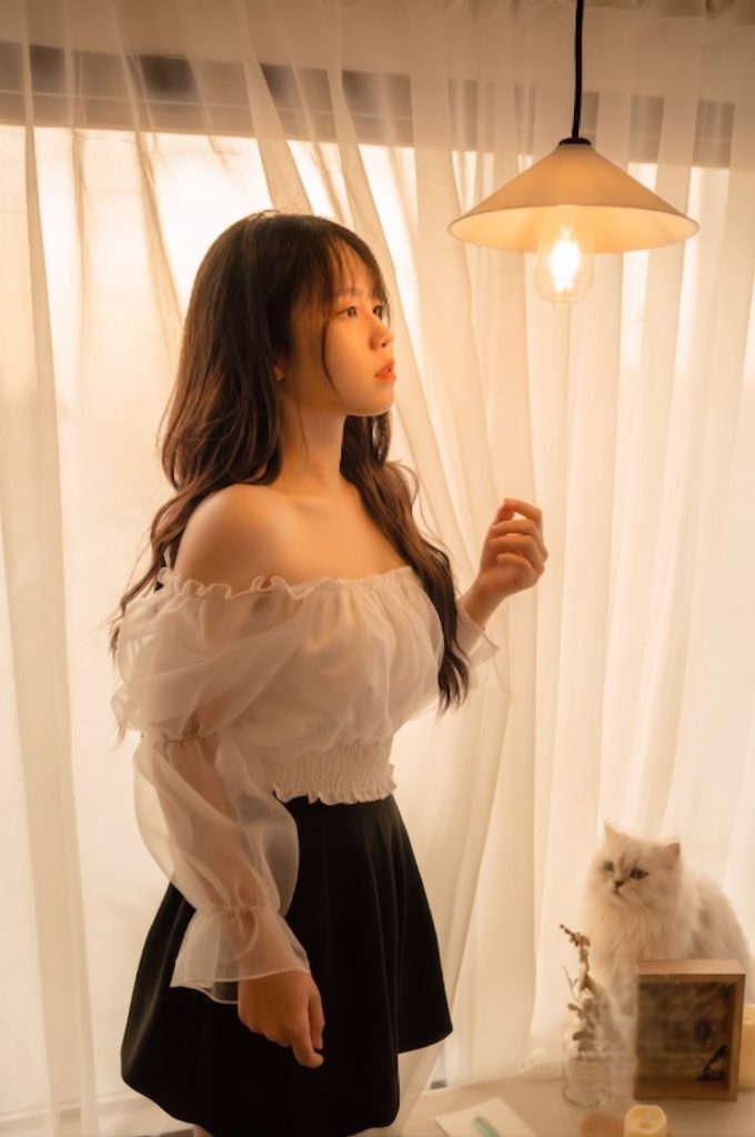 Young woman wearing a white off-the-shoulder sheer top standing near warm window curtains with a fluffy white cat nearby.