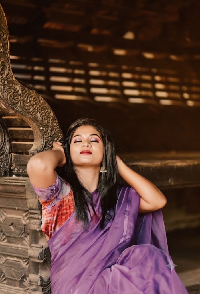 Woman with eyes closed wearing a purple saree and tie-dye blouse leaning against carved wooden architecture.