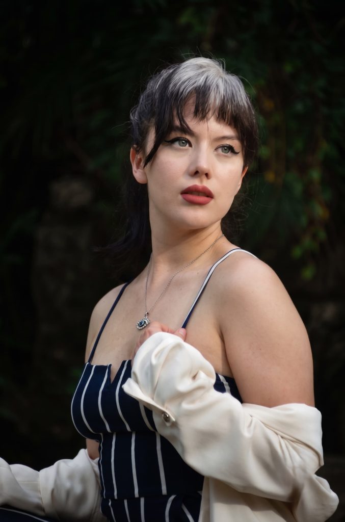 Woman with dark hair and silver-streaked bangs wearing a blue striped top and off-the-shoulder jacket.
