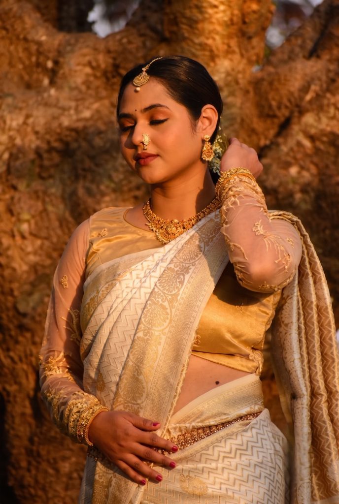 South Asian woman with eyes closed wearing an off-white and gold saree with traditional gold jewelry under warm sunlight.